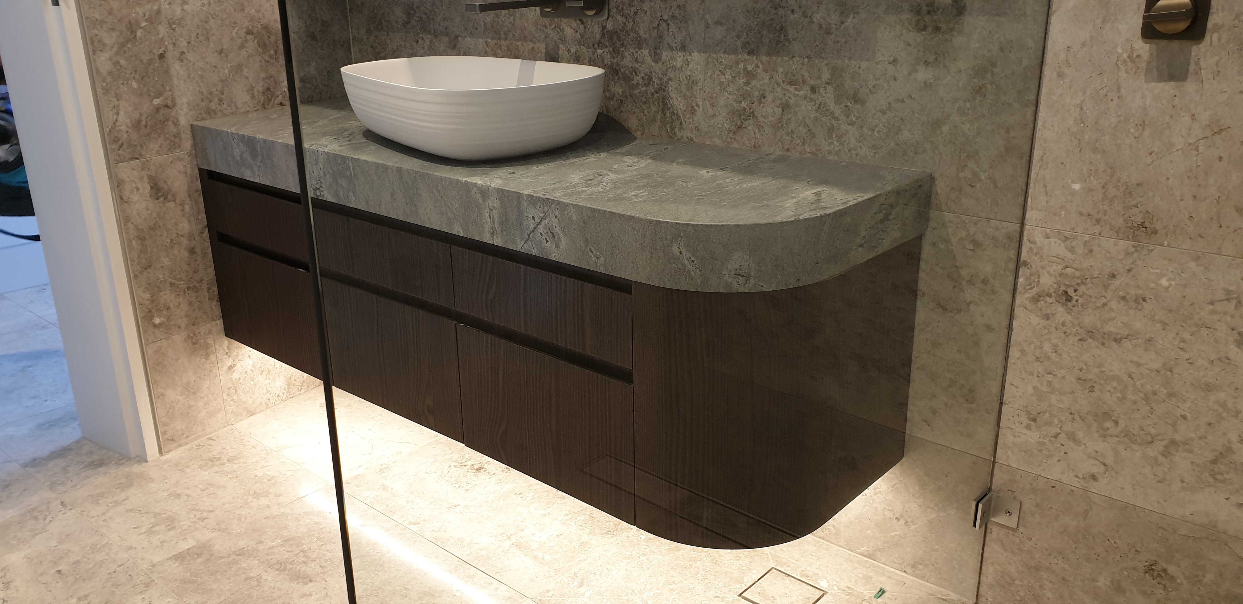 Curved grey stone vanity with a vessel basin and dark timber drawers